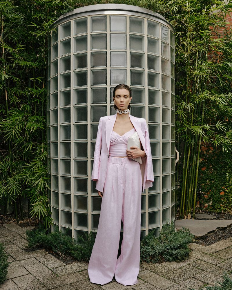 Fashionable woman in light pink suit poses outdoors against a glass block wall, surrounded by greenery and foliage in soft, natural lighting.
