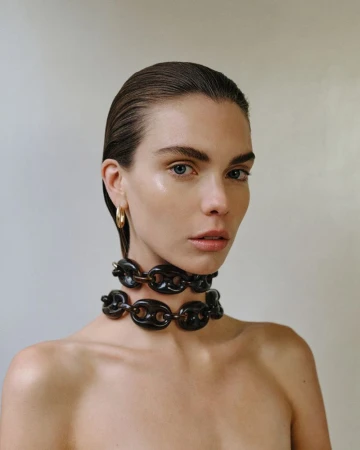 Close-up studio shot of a fair-skinned woman with wet slicked-back hair wearing a dark chain-link necklace and gold hoop earrings.