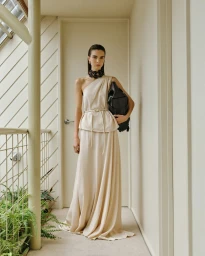 Model in a one-shoulder cream maxi dress with a black choker necklace holding a black bag in a sunlit neutral hallway.