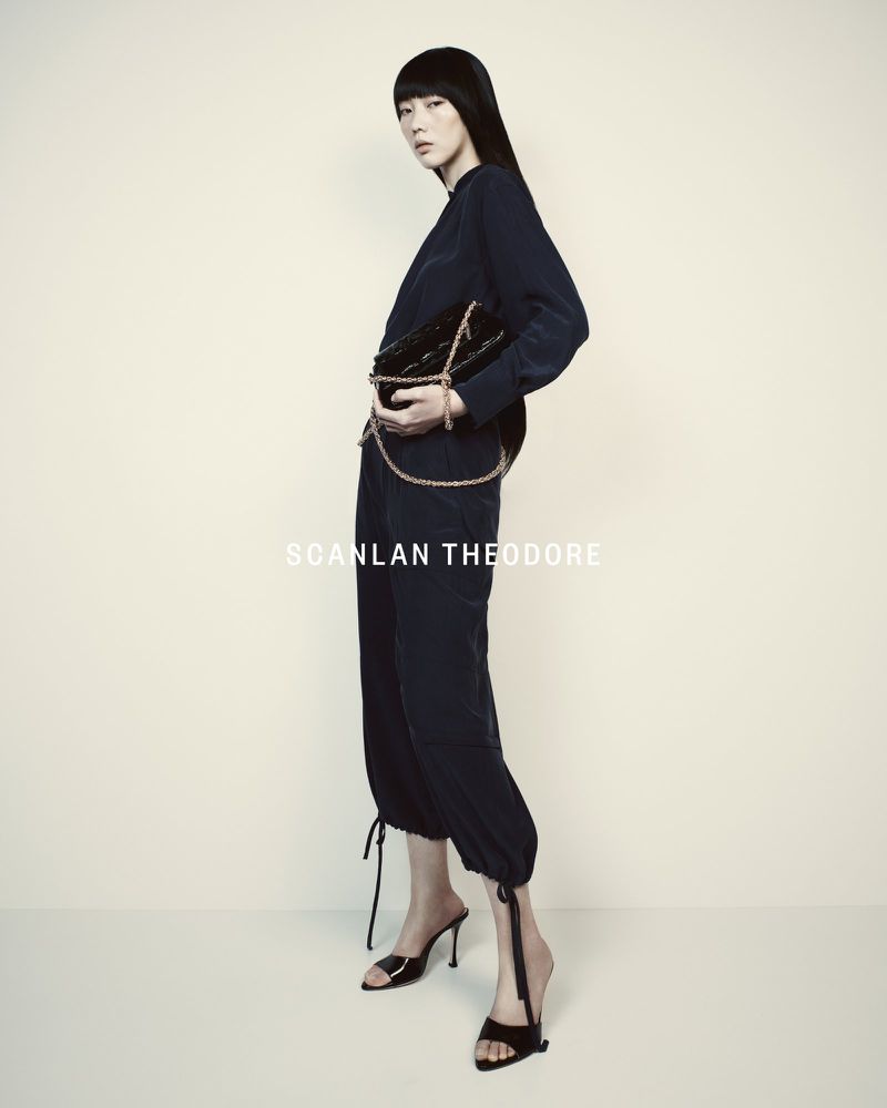 Fashionable adult female posing indoors wearing a navy outfit, black high-heel shoes, and holding a black quilted purse with gold chain.