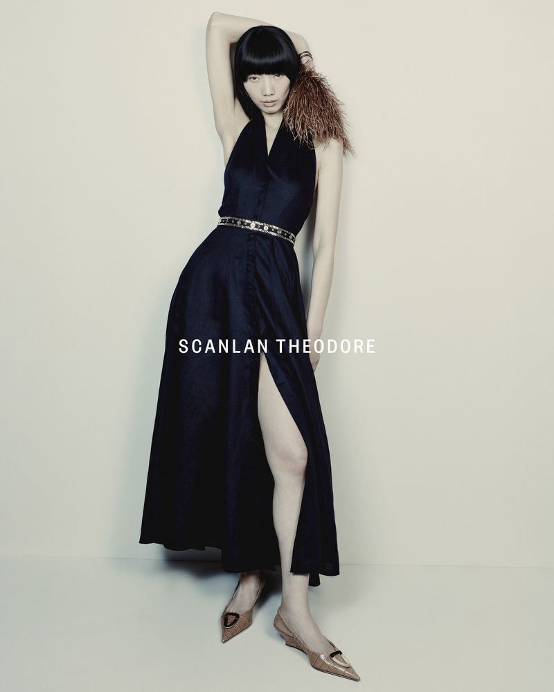 Elegant woman poses in a black halter dress with a thigh-high slit, accessorized with a belt and feathered shoulder detail in a studio setting.