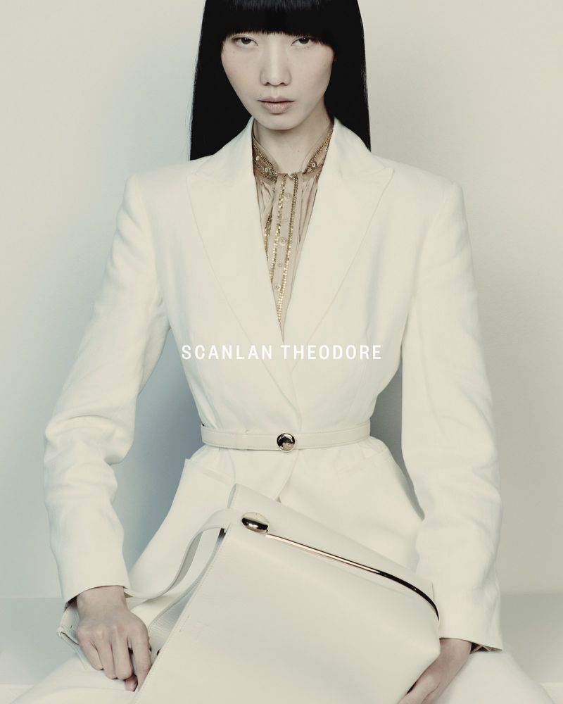 Fashion shot of an Asian woman in a white suit holding a white bag, posing against a light background with centered framing and neutral lighting.