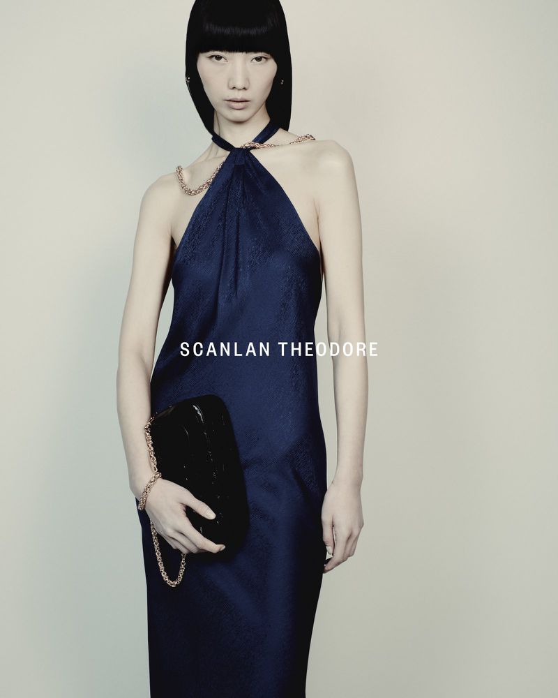 A sophisticated Asian woman presents a navy blue dress with chain detail, complemented by a shiny black clutch against a muted backdrop.