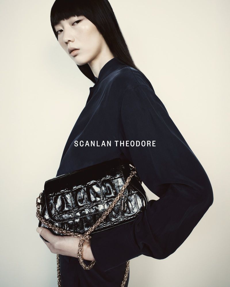 Fashion shoot featuring an adult female model holding a black purse with gold chain against a neutral studio backdrop, clothing by Scanlan Theodore.