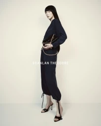 Model Yixin Zhao posing in a dark navy jumpsuit and black heels holding a chain strap clutch against an off-white background.