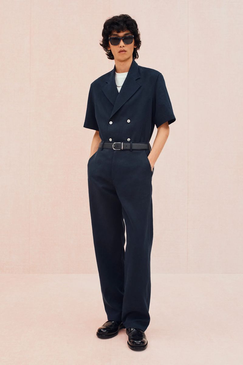 Full-length shot of an adult male in a navy short-sleeved double-breasted shirt, matching slacks, sunglasses, and black dress shoes, set against a peach backdrop.