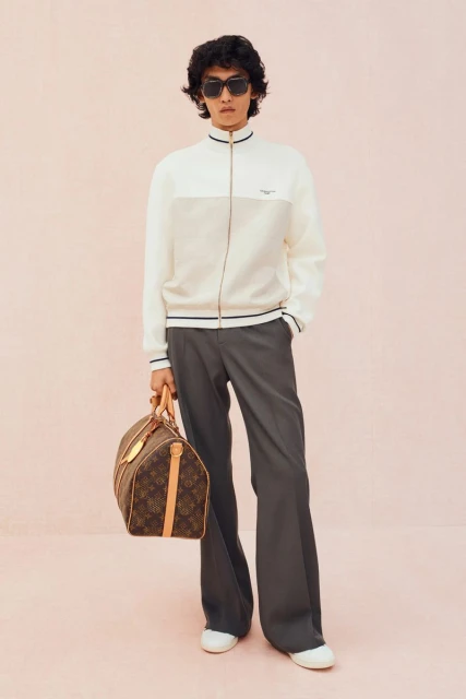 Male fashion model in a cream color-block zip jacket, grey trousers, and sunglasses holds a brown leather duffel bag against a pink background.