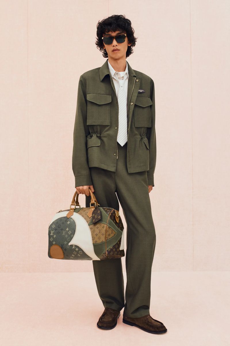 Man wearing a green jacket and pants, holding a patterned bag, posing against a light pink background in a studio setting.