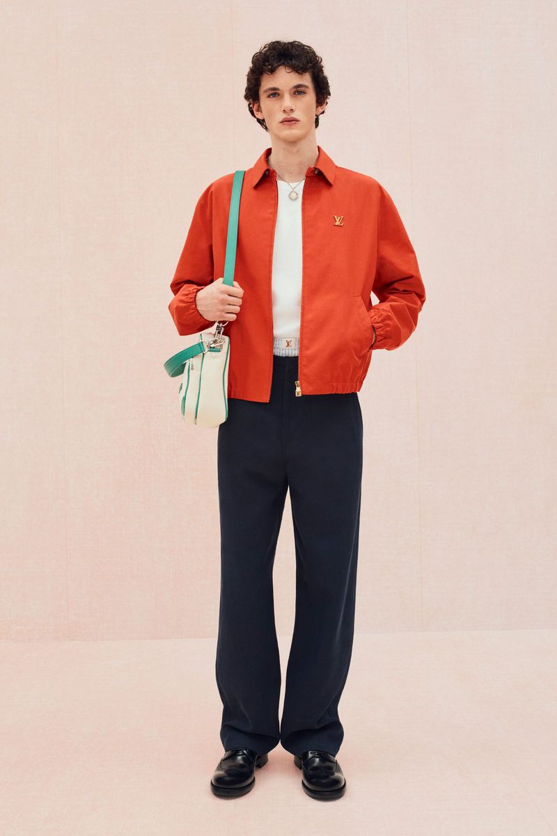 A young adult male poses in a full shot, wearing a red jacket, white shirt, navy pants, and black shoes with a shoulder bag.