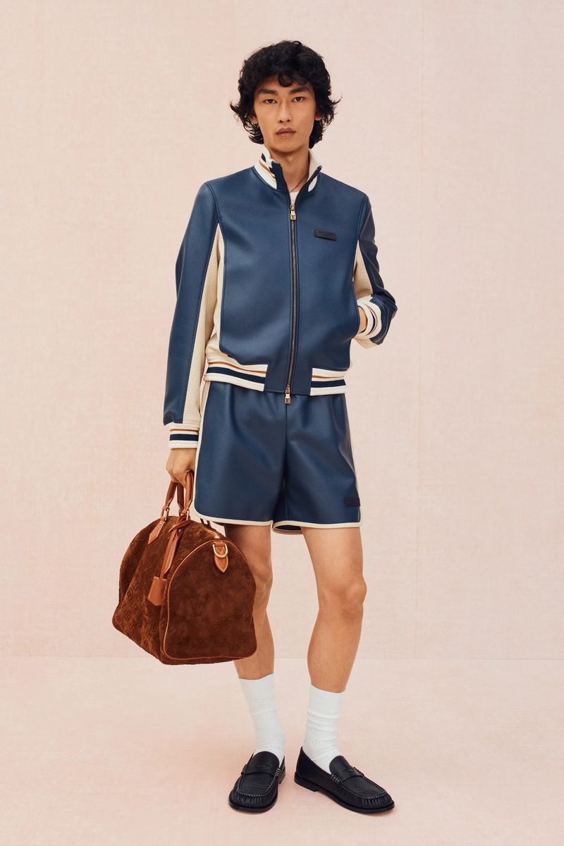 Asian male fashion model poses in matching blue leather jacket and shorts, holding brown suede duffel bag in studio setting.