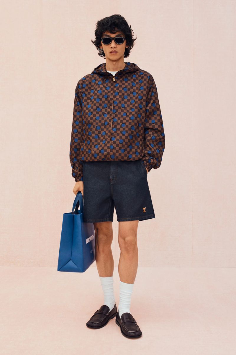 Man in patterned jacket and shorts holds blue Louis Vuitton tote bag against a pale pink backdrop, wearing sunglasses and loafers.