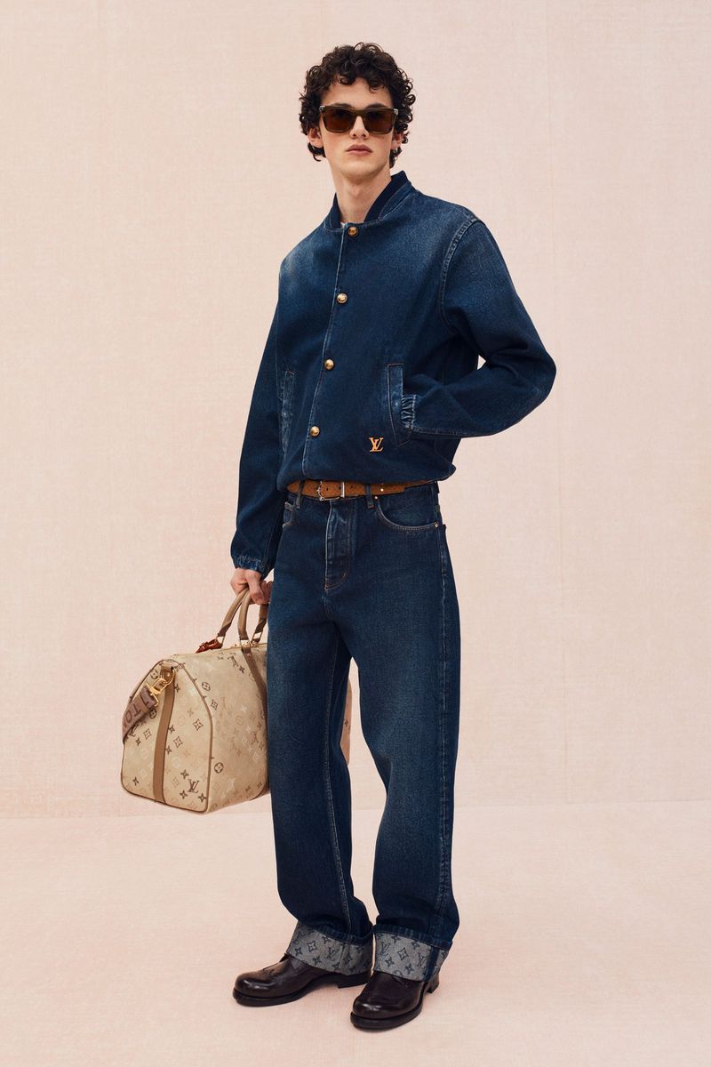 Man poses in denim jacket and jeans, holding patterned duffel bag against a pale backdrop in a full shot, fashionable and modern.