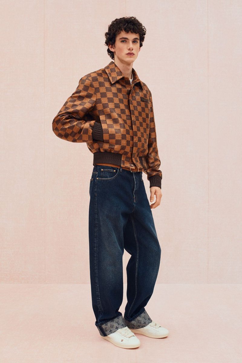 Fashion shot of a young adult male wearing a brown checkered bomber jacket with jeans and sneakers against a plain pink background.