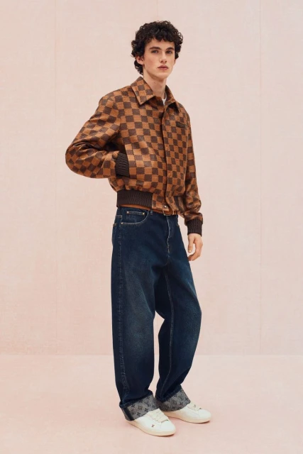 Fashion shot of a young adult male wearing a brown checkered bomber jacket with jeans and sneakers against a plain pink background.