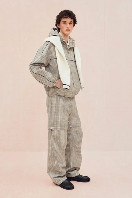 Full-shot of a young adult male model wearing a patterned tracksuit, with a sweater draped over his shoulders, against a plain backdrop.