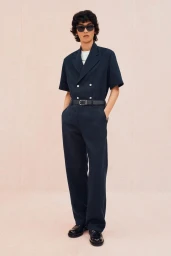 Model in dark sunglasses and a navy short-sleeve double-breasted jumpsuit posing against a pale pink studio backdrop.