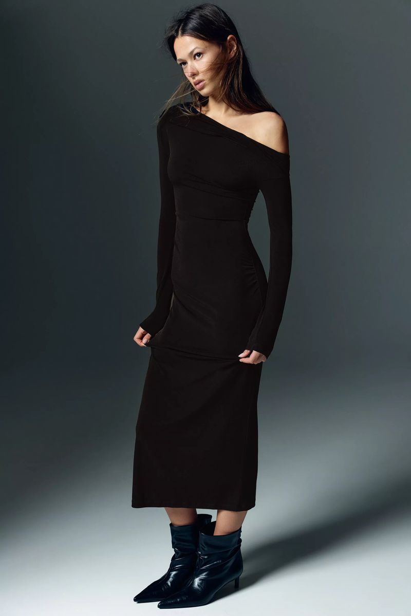 Brunette woman poses in a dark one-shoulder dress and black ankle boots against a grey gradient background in a fashion studio shot.
