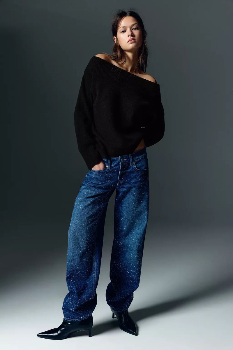 A young adult woman in a black sweater and blue jeans poses indoors against a grey background in a fashion shot.
