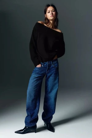 A young adult woman in a black sweater and blue jeans poses indoors against a grey background in a fashion shot.