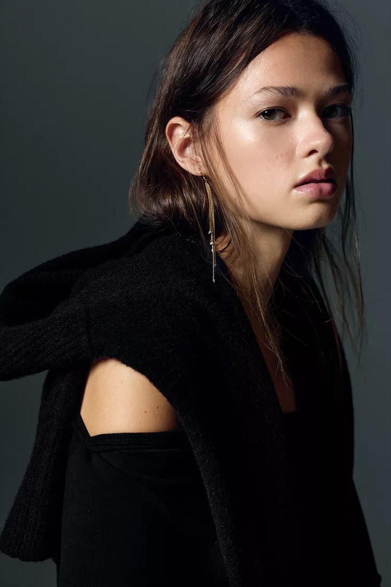 Close-up portrait of a young woman with long brown hair wearing a black top and sweater, adorned with a long, dangling earring.