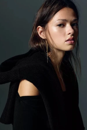 Close-up portrait of a young woman with long brown hair wearing a black top and sweater, adorned with a long, dangling earring.