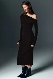 Model Didi Sviridova poses wearing an off-the-shoulder long-sleeve black dress and pointed black boots against a grey studio backdrop.