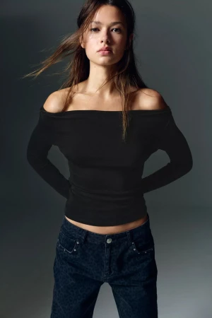 Model wearing an off-the-shoulder black top and dark jeans against a neutral grey studio background.