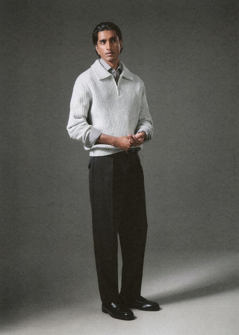 Adult male in fashion shot. Man with dark hair poses in collared sweater, dress pants, and shoes against a neutral backdrop.