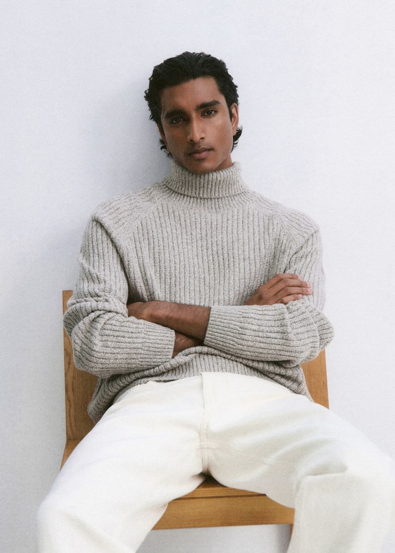 Model Jeenu Mahadevan seated in a wooden chair wearing a grey ribbed turtleneck sweater and white trousers against a white background.