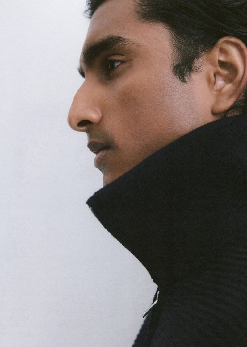 Portrait of an adult male of Indian descent in a close-up side view with a dark knit collar, against a plain light background in a studio.