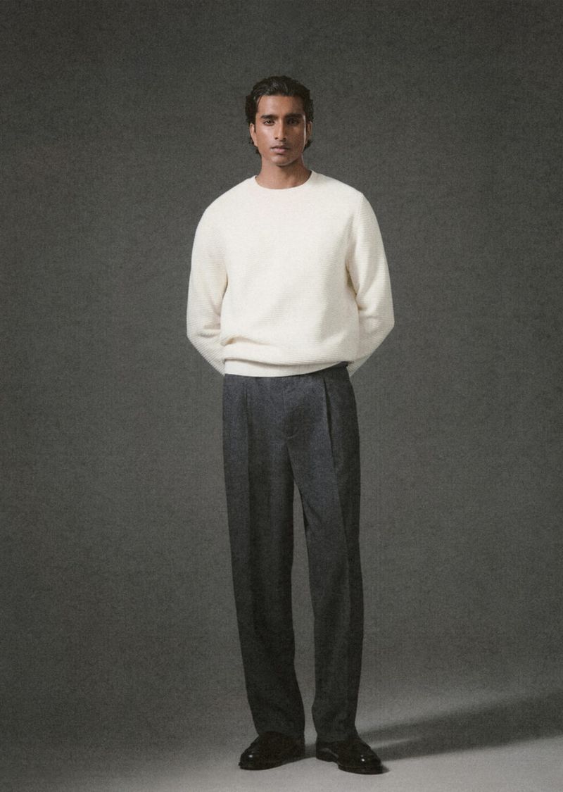 Full-body shot of a man with dark hair standing in a photo studio wearing a white sweater, grey wool pants, and black leather shoes.