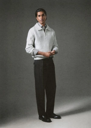 Model Jeenu Mahadevan in a light grey ribbed knit sweater and black trousers posing against a grey studio backdrop.