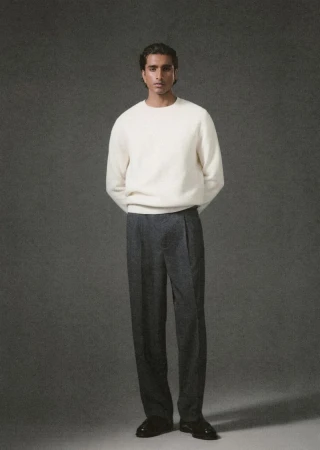 Man wearing a white knit sweater and grey trousers posing against a dark textured studio background.