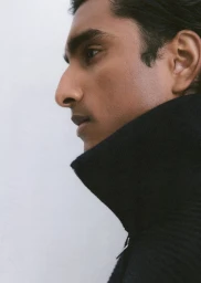 Profile close-up of model Jeenu Mahadevan wearing a high-collar black knit sweater against a plain pale backdrop.