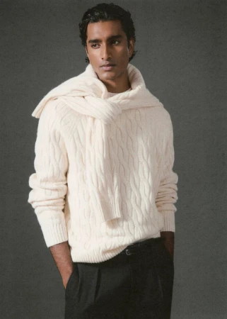 Jeenu Mahadevan wearing a cream cable-knit sweater with a matching knit draped over his shoulders and black trousers against a grey backdrop.