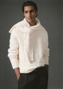 Jeenu Mahadevan wearing a cream cable-knit sweater with a matching knit draped over his shoulders and black trousers against a grey backdrop.
