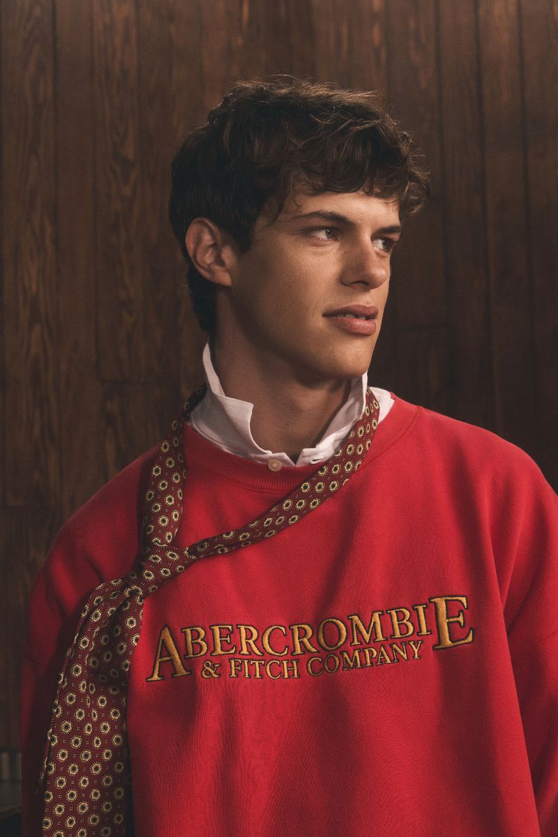 Close-up of a young adult male in a red Abercrombie & Fitch sweatshirt with a patterned tie, standing indoors against a wood-paneled wall.