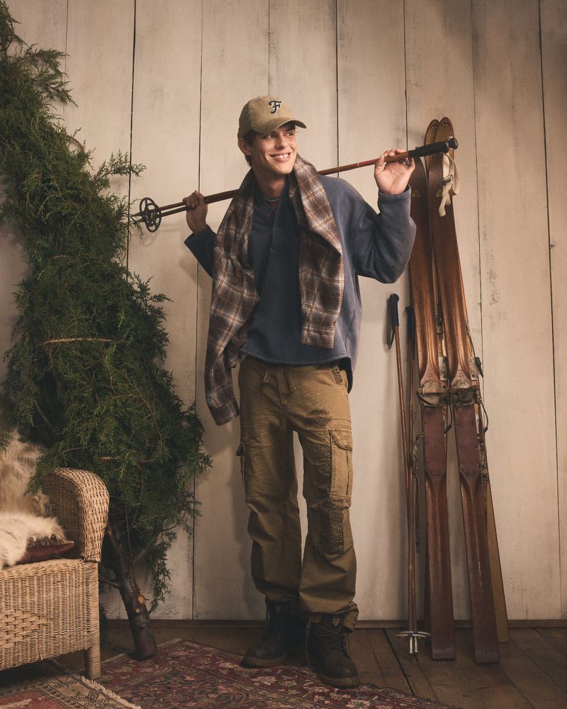 Man in cap and scarf with skis over his shoulder poses indoors with a Christmas tree and wicker chair on a patterned rug.