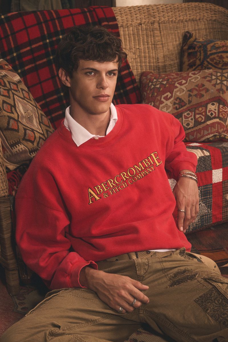 A male fashion model poses while wearing a red Abercrombie & Fitch Company sweatshirt and khaki cargo pants on a patterned settee.