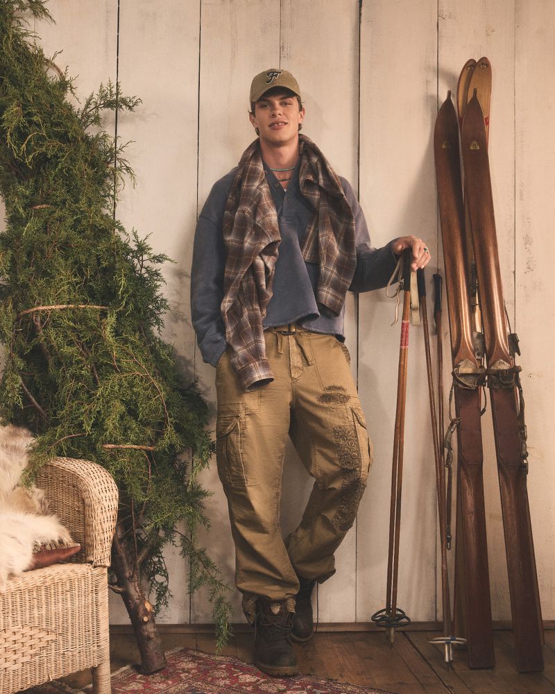 Man in cap and plaid scarf poses indoors with vintage skis. He wears a long-sleeve shirt and cargo pants, beside a wicker chair.