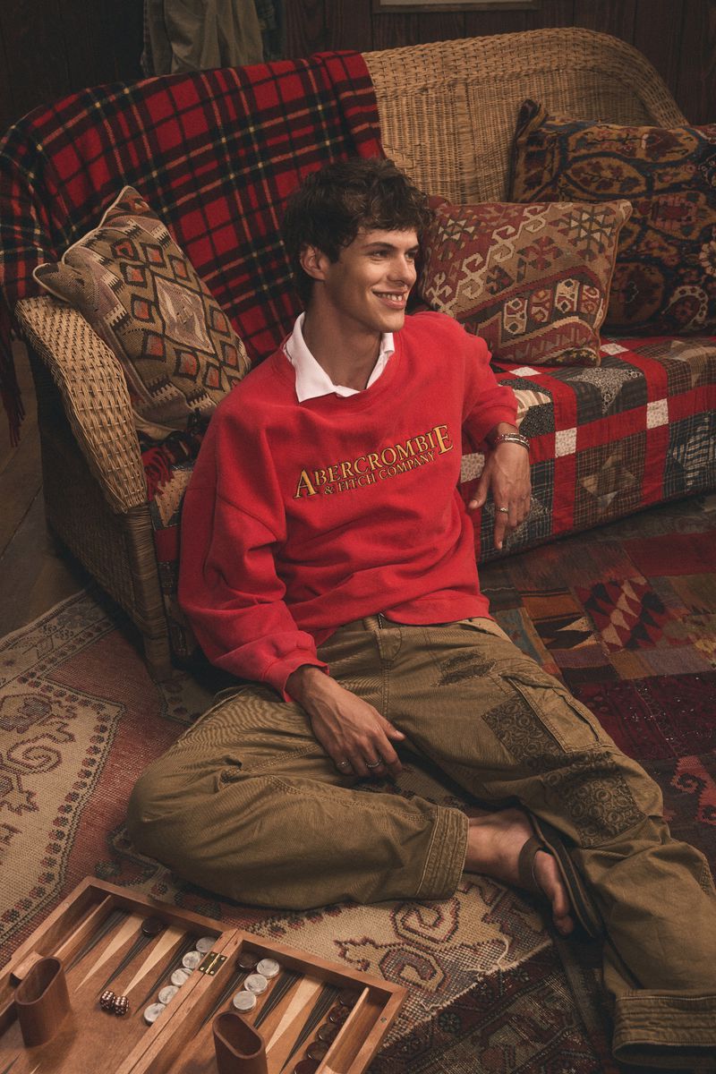Smiling man in red Abercrombie sweatshirt sits cross-legged on patterned rug with backgammon set in a casual indoor setting.