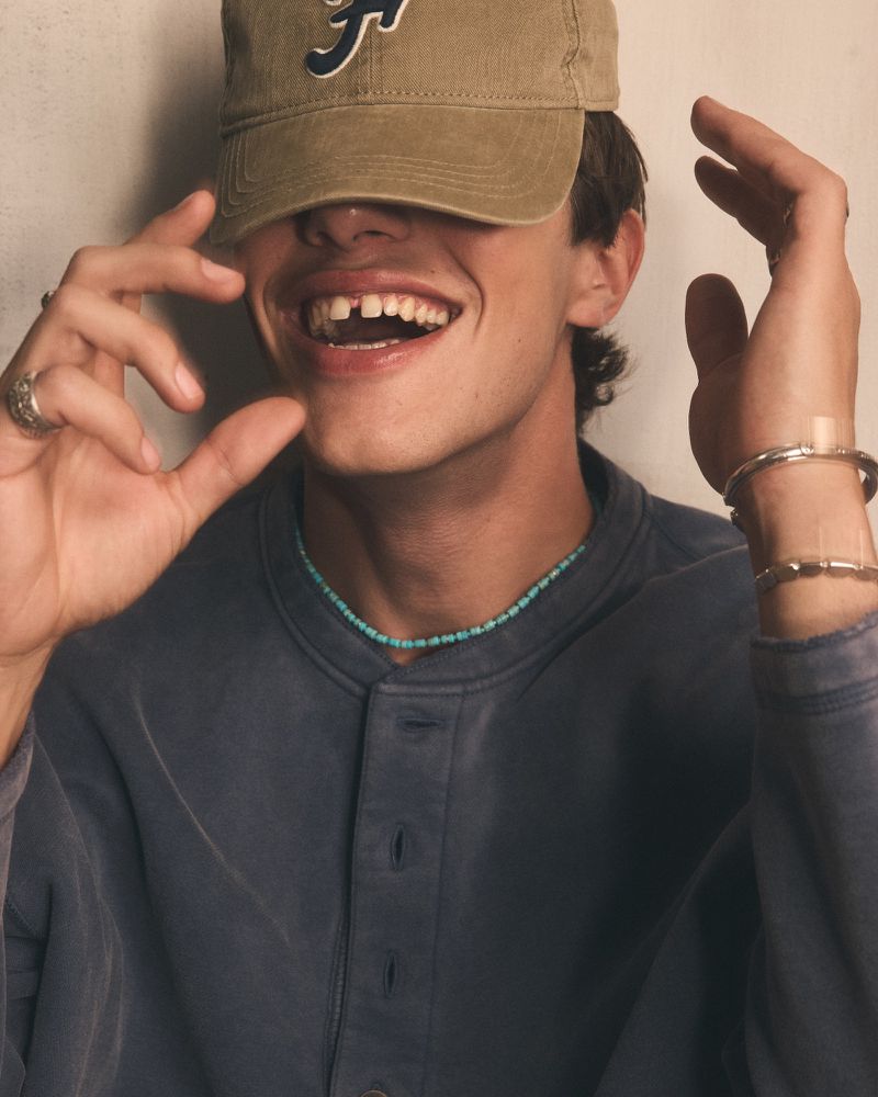 Smiling young man with missing tooth, wearing a cap, silver bracelets, and a beaded necklace against an off-white background.