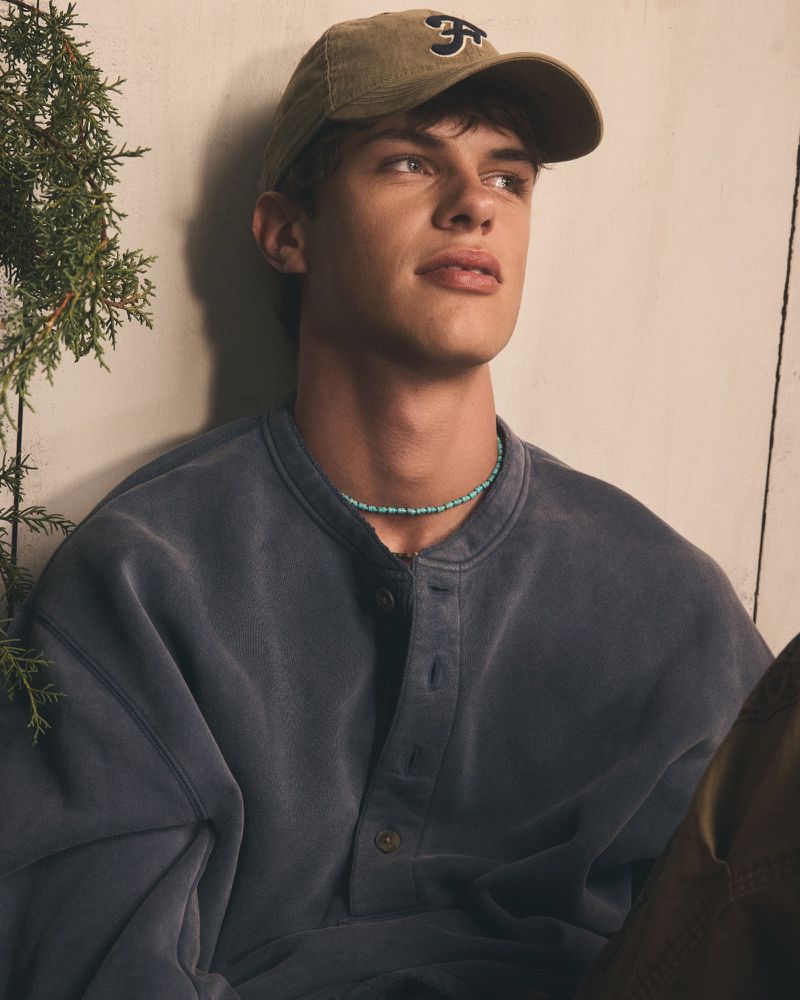 A young adult man wearing a baseball cap and sweatshirt, posing in a studio, with an evergreen branch to the side.