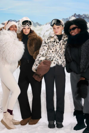 Group of women in snow wearing winter outerwear, goggles, and boots, posing in front of snow-covered mountains on a sunny day.