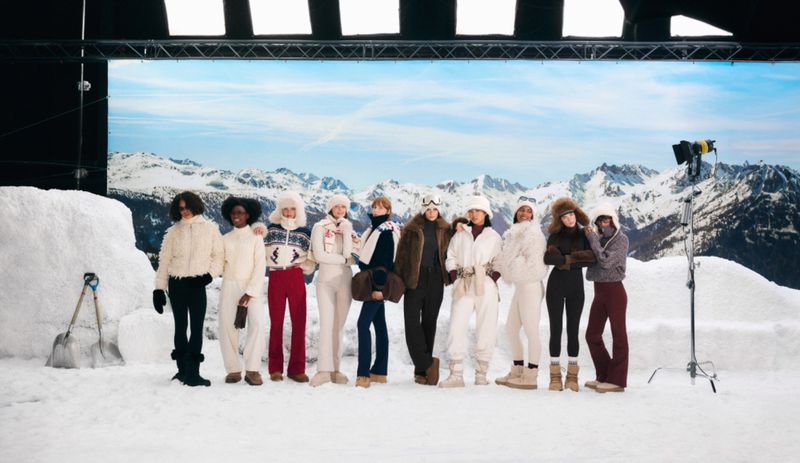 A diverse group of nine fashion models wearing winter ski clothes posing in a staged snowy photo studio with a mountain backdrop.