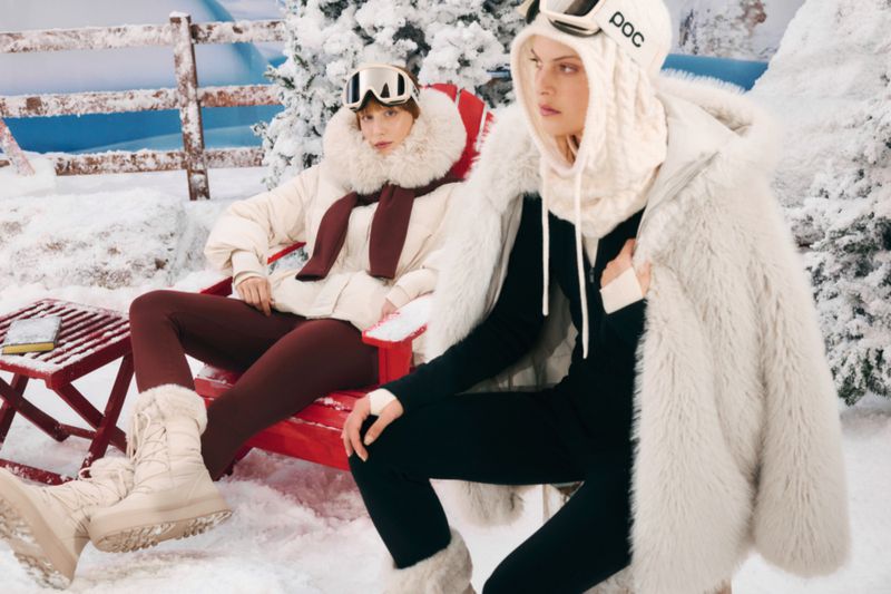Two women in winter ski fashion wearing goggles and warm clothing sitting in red chairs on snow with a winter scene backdrop.