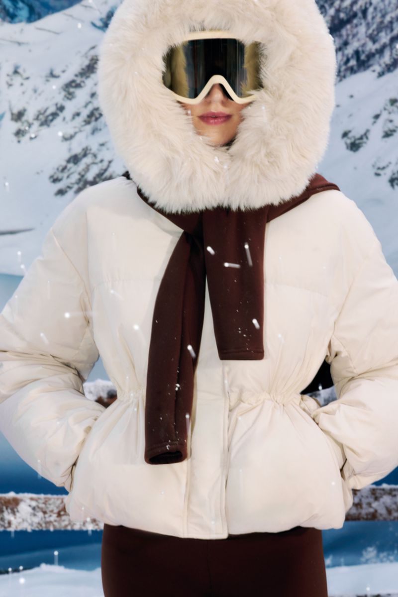 Adult female wearing a white puffer jacket with a fur-trimmed hood and brown scarf and leggings against a snowy mountain backdrop.