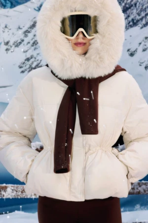 Adult female wearing a white puffer jacket with a fur-trimmed hood and brown scarf and leggings against a snowy mountain backdrop.