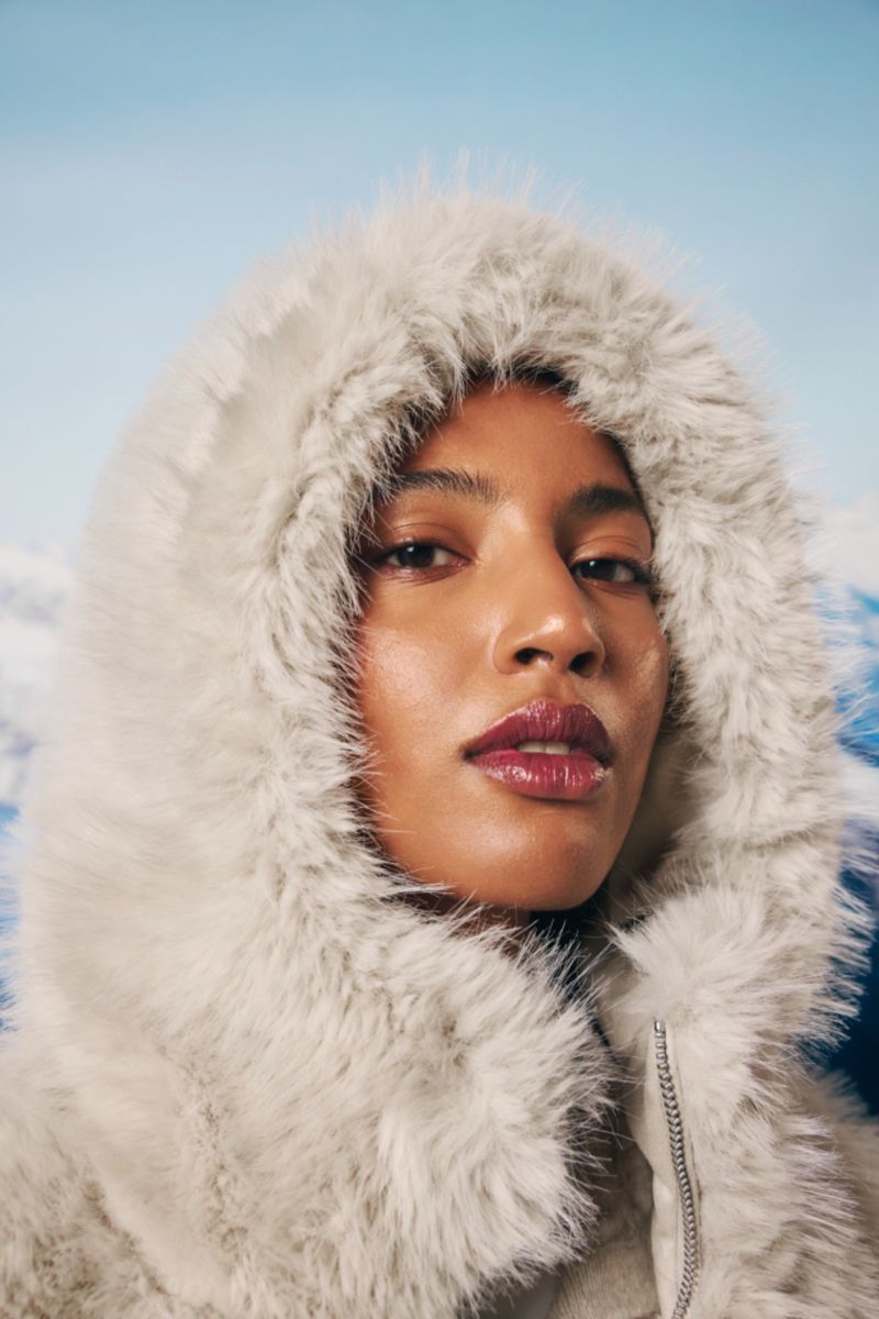 Close-up of a woman in a fur-trimmed hooded coat against a blurred blue sky and mountain background, fashion, winter style.