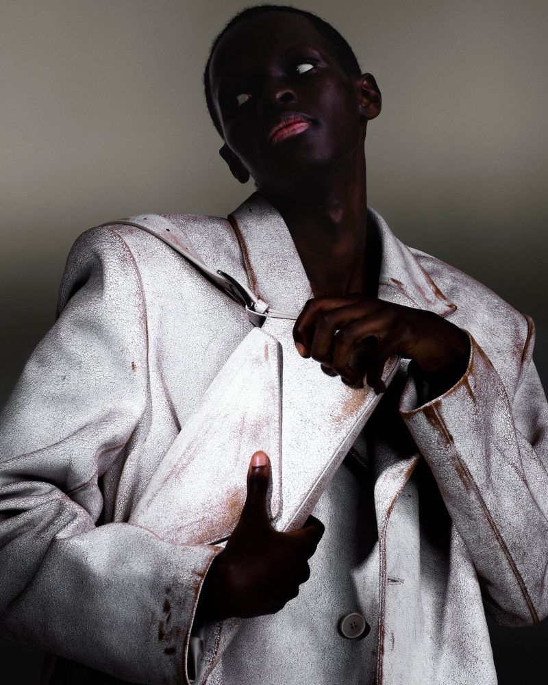 Dark-skinned model in a textured white jacket poses with a clutch bag, gazing up against a neutral backdrop in a fashion editorial.